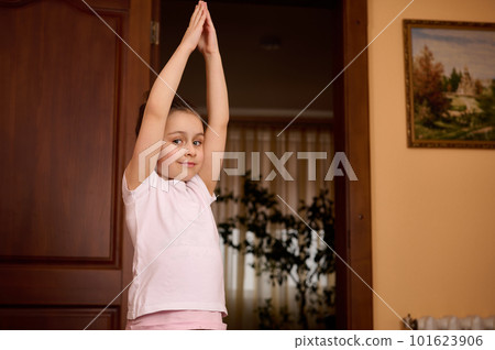 Caucasian beautiful little kid girl raising her arms up, stretching her body, practicing yoga, warming up while exercising indoors, smiling cutely, looking at camera. Waist-up portrait 101623906