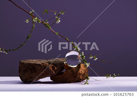 Front view of stone with transparent crystal ball, small flower branch on purple background. 101625763