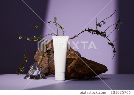 Front view of white plastic tube with facial moisturizer cream or facial cleanser on purple background. Brown stone with white flower as a highlight. 101625765