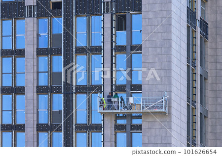 Builders on a suspended cradle mount decorative panels on the facade of a multi-storey skyscraper. Builders on a suspended cradle mount decorative panels on the facade of a multi-storey skyscraper. 101626654