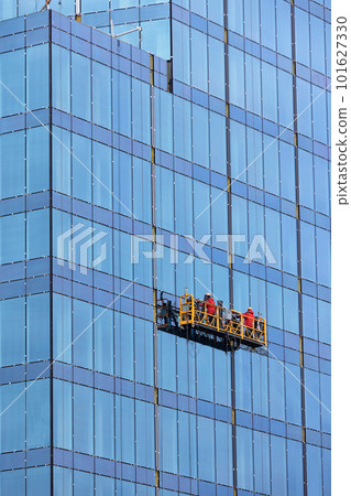 Builders in red overalls are mounting the glass facade of a multi-storey building on a suspended cradle. Builders in red overalls are mounting the glass facade of a multi-storey building on a suspended cradle. 101627330