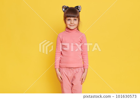 European adorable little female child posing isolated over yellow background, keeps hands at seams, looks at camera with calm expression, wearing pink pants and shirt. 101630573