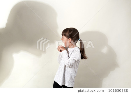 Domestic violence, abusing. Profile of scared little caucasian girl, victim child standing near gray wall with shadow of angry human threatening on it. Awareness of social problem, childhood. 101630588