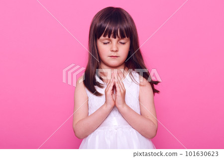 Calm beautiful little girl in white dress practicing yoga, keeping eyes closed, pressing palms together, dark haired cute kid praying, expressing hope, isolated over pink background. Calm beautiful little girl in white dress practicing yoga, keeping eyes closed, pressing palms together, dark haired cute kid praying, expressing hope, isolated over pink background. 101630623