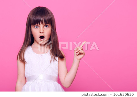 Surprised female child wearing beautiful white dress pointing aside with fore finger with widely opened mouth, sees something astonishing, copy space, isolated over pink background. 101630630