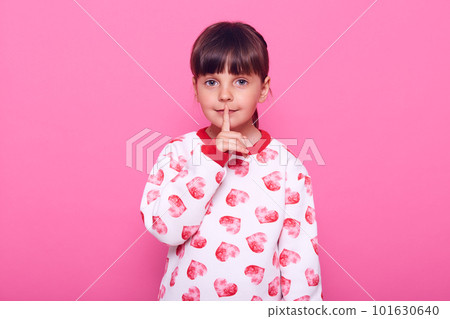Little girl of preschool age, holds her finger near her lips, shows that she would not tell the secret, dresses in stylish sweater, has dark hair, isolated over pink background. 101630640