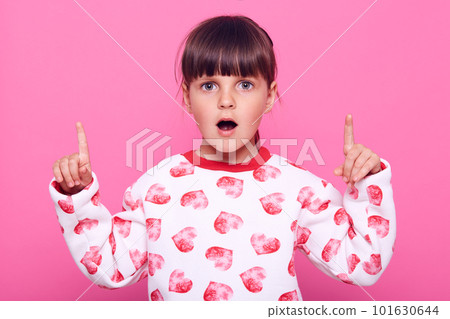 Charming surprised female child in casual sweater pointing with both index fingers up, advertising shocking offers, looks at camera with astonished expression, isolated over pink background. 101630644