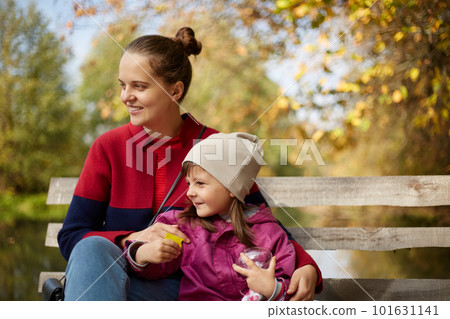 Young dark haired mother with daughter sitting on wooden bench in autumn park near lake or river, family having rest by water, mom and little girl looking in distance and enjoying nature. Young dark haired mother with daughter sitting on wooden bench in autumn park near lake or river, family having rest by water, mom and little girl looking in distance and enjoying nature. 101631141