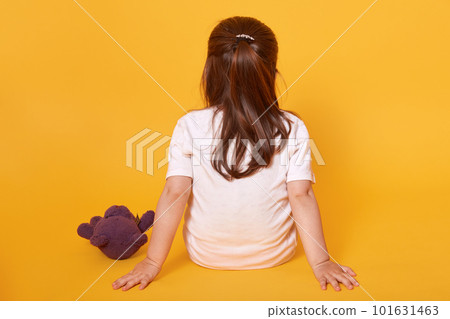 Rear view of little fair haired child sitting on the floor, putting hands aback, being punished, spending time alone, having brown teddy bear nearby, wearing white t shirt. Childhood concept. 101631463