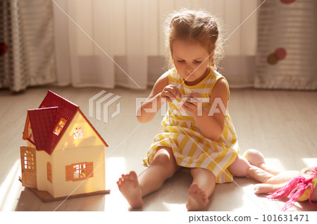 Horizontal shot of little girl playing with wooden doll house, funny female child wearing yellow stripped dress enjoys playing with her favourite toy in children's room, repeairing her small girl. Horizontal shot of little girl playing with wooden doll house, funny female child wearing yellow stripped dress enjoys playing with her favourite toy in children's room, repeairing her small girl. 101631471