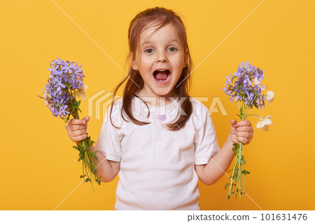 Picture of emotional playful little girl opening mouth and eyes widely, holding flowers in both hands, wearing white t shirt, preparing present for her mom on women day. Children and gift concept. 101631476