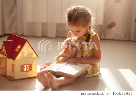 Little charming gir with pigtails reading her favorite book for her soft toy. Cute child reads stories, sitting on floor near toy house. Self isolation during coronavirus pandemic. Covid 19 concept. 101631485