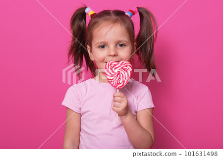 Young pretty toddler girl kid with big sweet lollypop candy in rose casual t shirt posing isolated over pink studio background, has two funny ponitails with colourful scrinchies. Childhood concept. Young pretty toddler girl kid with big sweet lollypop candy in rose casual t shirt posing isolated over pink studio background, has two funny ponitails with colourful scrinchies. Childhood concept. 101631498