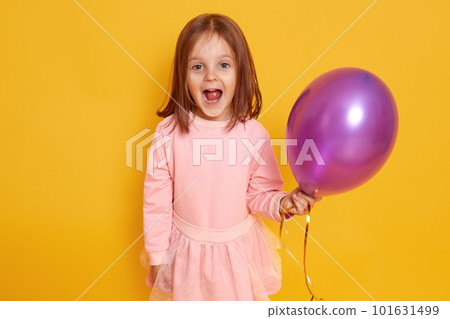 Portrait of surprised little girl with dark staright hair standing over yellow studio background beautiful clothes, holding purple ballon in hands and looking at camera joyfully. Holiday concept. Portrait of surprised little girl with dark staright hair standing over yellow studio background beautiful clothes, holding purple ballon in hands and looking at camera joyfully. Holiday concept. 101631499
