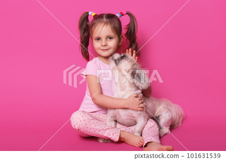 Studio shot of little funny child sitting on floor, looking directly at camera, hugging her pet, dog enjoying time with kid, sweet girl wearing rose suit, having pony tails. Children and pets concept. Studio shot of little funny child sitting on floor, looking directly at camera, hugging her pet, dog enjoying time with kid, sweet girl wearing rose suit, having pony tails. Children and pets concept. 101631539