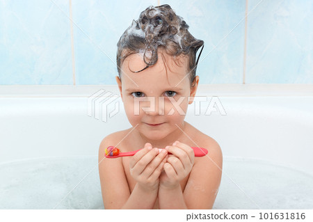 Portrait ot attractive little child looking at camera, keeps red toothbrush in hands, ready for brushing teeth, isolated over blue wall in bathroom, being glad to take bath and photographed. 101631816