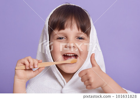 Close up portrait of funny pleasant happy little girl making gesture, showing sign super, thumb up, having towel on head, brushing teeth, looking directly at camera. Childhood and emotions concept. 101631828