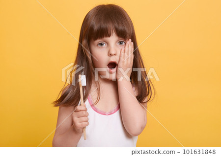 Studio shot of emotional impressed little girl opening mouth widely, having surprised facial expression, putting hand on cheeck, holding toothbrush, looking directly at camera. Hygiene concept. 101631848