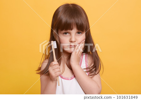 Closeup portrait of serious cute child keeping palm on cheek, having toothache, holding toothbrush in hand, looking at camera, standing isolated over yellow background. Daily dental hygiene concept. Closeup portrait of serious cute child keeping palm on cheek, having toothache, holding toothbrush in hand, looking at camera, standing isolated over yellow background. Daily dental hygiene concept. 101631849