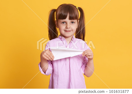 Image of delighted positive girl standing isolated over yellow background, holding paper plane in both hands, having fun, playing game, looking directly at camera, having peaceful facial expression. Image of delighted positive girl standing isolated over yellow background, holding paper plane in both hands, having fun, playing game, looking directly at camera, having peaceful facial expression. 101631877
