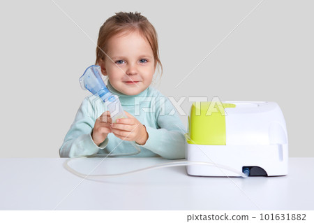 Small attractive girl makes inhalation with nebulizer, cures cough, sits at white desk against studio background. Compressor inhaler with mask. Little kid has allergy. Medicine and health concept Small attractive girl makes inhalation with nebulizer, cures cough, sits at white desk against studio background. Compressor inhaler with mask. Little kid has allergy. Medicine and health concept 101631882