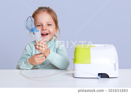 Little girl making inhalation with nebulizer at home, child suffers from asthma, uses inhaler, sits at white desktop, looks smiling at camera, holds mask in hand. Charming kid cures cough or pneumonia 101631883