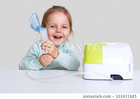 Photo of small child laughs happily, does inhalation, cures asthma, dressed in light blue turle neck sweater, isolated over white background. Therapy with inhaler. Kid with respiratory problem 101631885