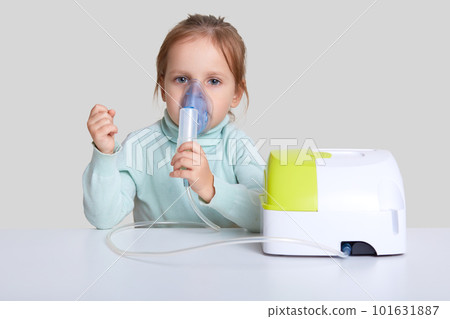 Beautiful sick girl has inhalation therapy, uses portable nebulizer, holds mask vapor, sits at white desktop, isolated over studio background. Respiratory disease. Shallow depth. Health concept 101631887