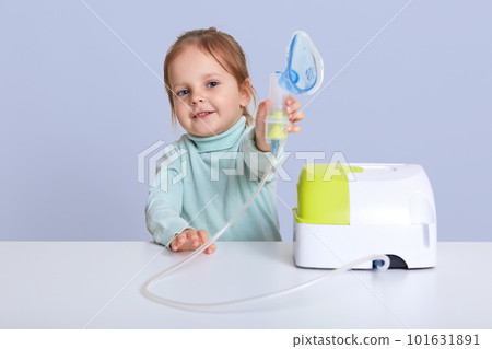 Small attractive girl shows inhaler, makes inhalation with nebulize, sits at white desk, treats illness, dressed casual sweater, posing against blue studio background. Children and health concept. Small attractive girl shows inhaler, makes inhalation with nebulize, sits at white desk, treats illness, dressed casual sweater, posing against blue studio background. Children and health concept. 101631891