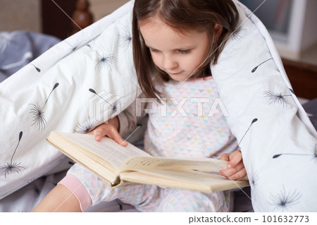 Horizontal shot of girl reading under blanket with dandelion, having serious facial expression, looks concentrated, charming toddler holding book in hands, reads fairy tale. Childhood concept. Horizontal shot of girl reading under blanket with dandelion, having serious facial expression, looks concentrated, charming toddler holding book in hands, reads fairy tale. Childhood concept. 101632773