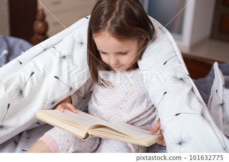 Portrait of charming child reading book at home, wrapped white blanket with dandelions, dark haired kid wearing pajama looking concentrated down on pages from her book. Children education concept. 101632775