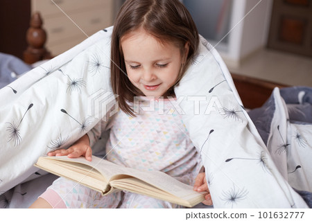 Horizontal shot of little girl reading under blanket with dandelion, having happy facial expression, looks exited, charming female child holding book in hands, reads fairy tale. Childhood concept. Horizontal shot of little girl reading under blanket with dandelion, having happy facial expression, looks exited, charming female child holding book in hands, reads fairy tale. Childhood concept. 101632777