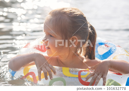 Outdoor shot of fair haired funny little girl spending free time in lake, learning to swim with swimming circle, looking aside, holding on to rubber circle, enjoying water activities. Rest concept. Outdoor shot of fair haired funny little girl spending free time in lake, learning to swim with swimming circle, looking aside, holding on to rubber circle, enjoying water activities. Rest concept. 101632778