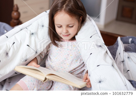 Horizontal indoor shot of playful smart sweet kid holding book in both hands, looking through attentively, sitting in her bedroom under blanket, wearing pyjamas. Children and free time concept. Horizontal indoor shot of playful smart sweet kid holding book in both hands, looking through attentively, sitting in her bedroom under blanket, wearing pyjamas. Children and free time concept. 101632779