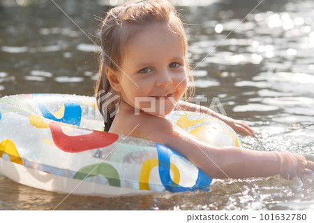 Close up portrait of playful cheerful kid smiling sincerely, having pleasant facial expression, looking directly at camera, swimming in river, holding on to swimming circle, being in high spirits. 101632780