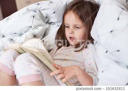Close up portrait of little girl reading story or fairy tale for her teddy bears while laying in bed and wearing pajama, charming kid speling words aloud, looks concentrated, being in good mood. Close up portrait of little girl reading story or fairy tale for her teddy bears while laying in bed and wearing pajama, charming kid speling words aloud, looks concentrated, being in good mood. 101632811