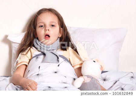 Charming cute child lying in her bed with toy, looking directly at camera, opening her mouth widely, being on sick leave, staying at home, feeling unwell, having sore throat and cough. Health concept. 101632849