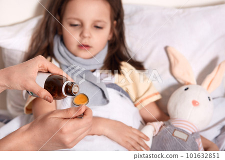 Horizontal photo of unknown hand pouring liquid into spoon from little bottle with syrup, person caring about child lying in bed with flu and sore throat, being upset. Children and health concept. 101632851