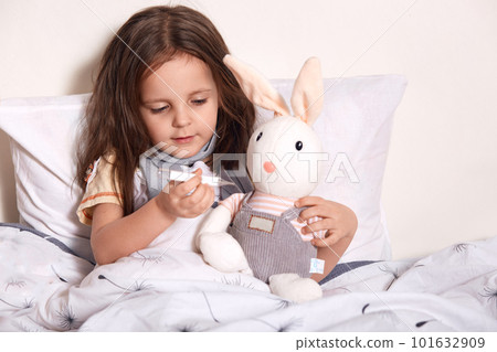 Little girl lying in bed and measuring soft hear's temperature with thermometer, child lying in bed on pillow with her favourite toy, wearing gray scarf around neck, suffering from flue, cathing cold. Little girl lying in bed and measuring soft hear's temperature with thermometer, child lying in bed on pillow with her favourite toy, wearing gray scarf around neck, suffering from flue, cathing cold. 101632909