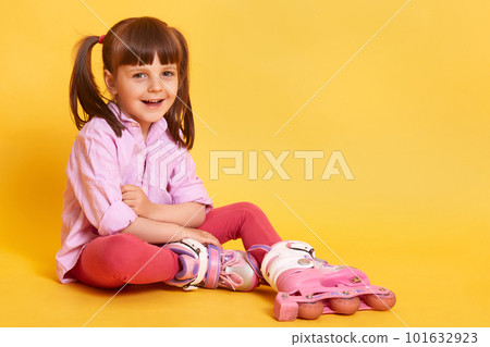 Studio shot of happy smiling girl sitting on floor, looks at camera, spending weekend in active way, having two ponytail, wearing casual attire, poses isolated on yellow background. Childhood concept. Studio shot of happy smiling girl sitting on floor, looks at camera, spending weekend in active way, having two ponytail, wearing casual attire, poses isolated on yellow background. Childhood concept. 101632923