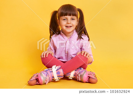 Horizontal shot of happy smiling girl wearing casually, having two ponytails, looks at camera, child spends leisure time rollerblading, being photographed while having rest. Children concept. 101632924