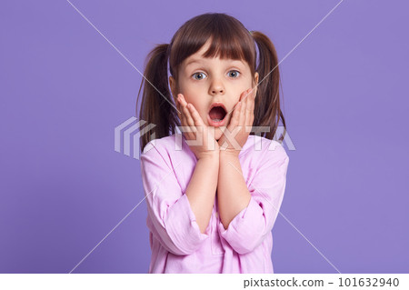 Studio shot of astonished female kid with widely opened mouth, keeping hands on cheeks, looks at camera with big eyes, standing against lilac background, child having surprised facial expression. Studio shot of astonished female kid with widely opened mouth, keeping hands on cheeks, looks at camera with big eyes, standing against lilac background, child having surprised facial expression. 101632940