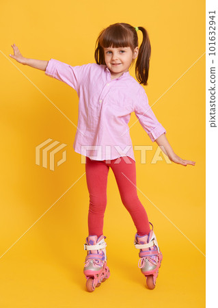 Pleasant looking kid raising hands while wearing roller skates, having two ponytails, standing isolated over yellow background, having fun and looks at camera. Pleasant looking kid raising hands while wearing roller skates, having two ponytails, standing isolated over yellow background, having fun and looks at camera. 101632941