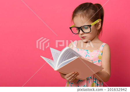 Image of cute preschooler girl wearing sundress and eyewear, female kid reading textbook while standing isolated over rose studio background, child enjoying to read interesting fairy tale. Education. 101632942