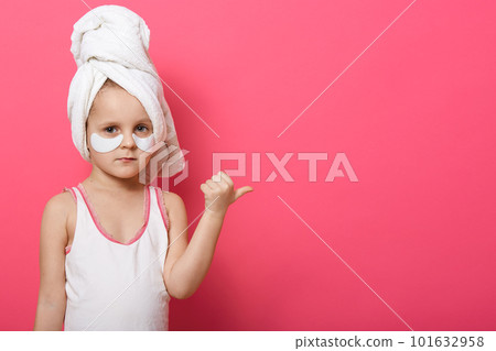 Close up portrait of female child wearing white towel on her head and having patches under eyes, kid with serious facial expression, points aside with thumb. Copy space for advertidsment or promotion. Close up portrait of female child wearing white towel on her head and having patches under eyes, kid with serious facial expression, points aside with thumb. Copy space for advertidsment or promotion. 101632958