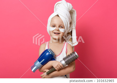 Horizontal shot of charming female kid posing isolated over rose studio background, little girl holding brush and hair dryer, kid making hairstyle, posing with towel on head and patches under eyes. 101632965