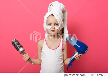 Portrait of cute good looking little child holding round hair brush and hair dryer in both hands, having patches under eyes, wearing pajama, having towel on head, taking care, hygiene and cleaning. Portrait of cute good looking little child holding round hair brush and hair dryer in both hands, having patches under eyes, wearing pajama, having towel on head, taking care, hygiene and cleaning. 101632972