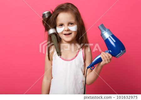 Studio shot of charming cute female kid posing isolated over rose studio background with blue hairdryer in hands, little girl with combin her hair, having tangled hairbrush on head. Children concept. Studio shot of charming cute female kid posing isolated over rose studio background with blue hairdryer in hands, little girl with combin her hair, having tangled hairbrush on head. Children concept. 101632975