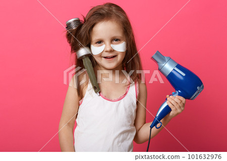 Close up portrait of beautiful little smiling girl with hair dryer and brush in hands, posing isolated over rosy studio wall with charming smile, having patches under eyes, looking at camera. Close up portrait of beautiful little smiling girl with hair dryer and brush in hands, posing isolated over rosy studio wall with charming smile, having patches under eyes, looking at camera. 101632976