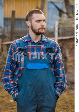 Farmer handsome european appearance male rural portrait with beard, shirt and overalls with hands in pockets looking at outdoors countryside 101637924